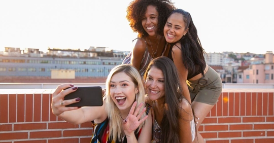 four women standing by a brick wall taking a photo on a phone